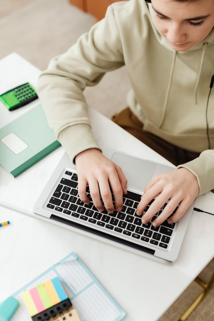 Teenager in hoodie using a laptop for online education with study materials around.
