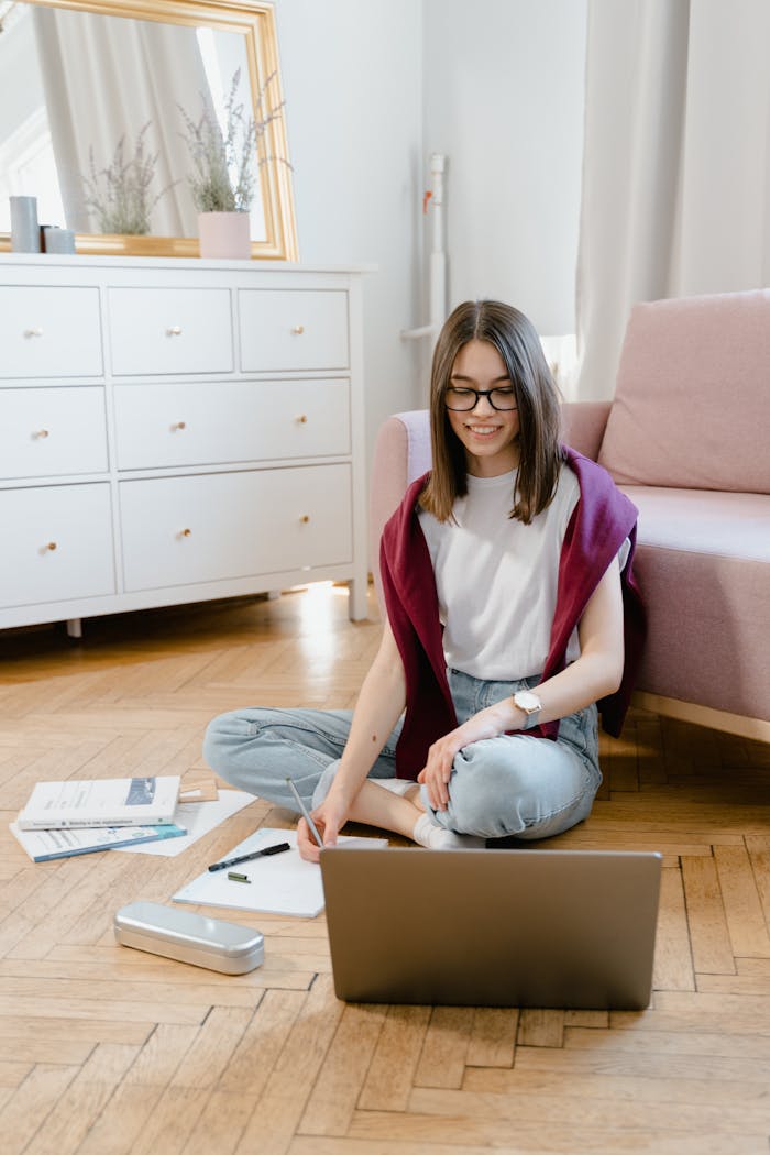 Teen girl engaged in online learning at home, writing notes with laptop on the floor.
